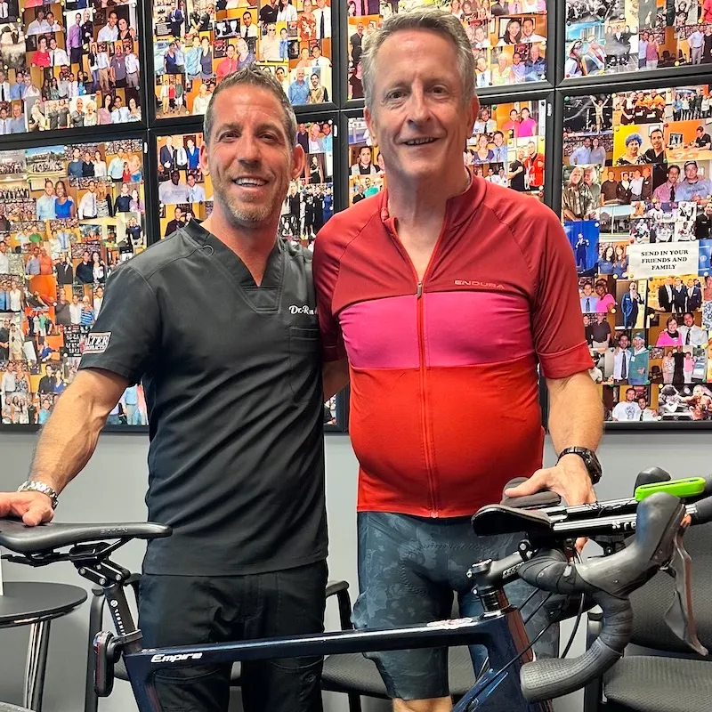 Dr Ryan Alter and a man stand beside a bicycle in front of a wall covered with photos